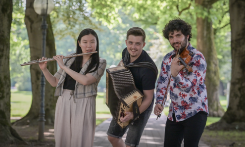 1 Frau Querflöte, 1 Mann Zieharmonika und einer Geige spielend blicken fröhlich in die Kamera