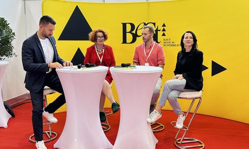 2 Frauen und 1 Mann sitzen auf Barhockern rund um 2 Stehtischen und sind im Gespräch